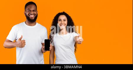 Multiethnic Couple Showing Cellphone Screen Gesturing Thumbs-Up Over Yellow Background Stock Photo