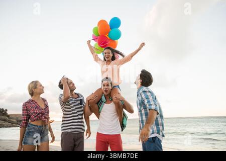 Caucasian young man carrying woman with balloons on shoulders while standing with friends at beach Stock Photo