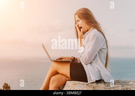 Woman Laptop Beach Surprised - A woman sits on a rock overlooking the ocean, using her laptop and reacting with surprise. Stock Photo
