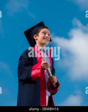 Handsome boy with elementary school graduation outfit and diploma. Boy in graduation robes looking high up with sky background Stock Photo