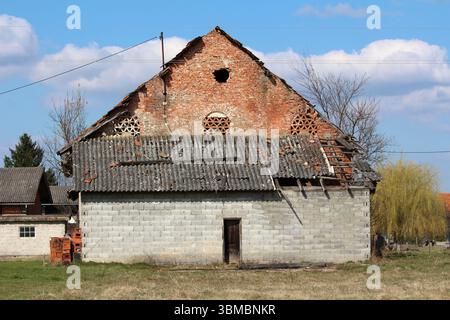 Old barn constructed of red brick and gray concrete blocks, featuring a partially collapsed tile roof and missing sections, stands in a grassy rural a Stock Photo