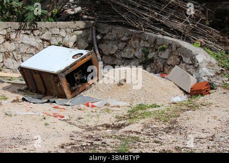 Rusted metal appliance lies on its side among gravel, bricks, and debris at a construction site, surrounded by stone wall, sand pile, and scattered Stock Photo