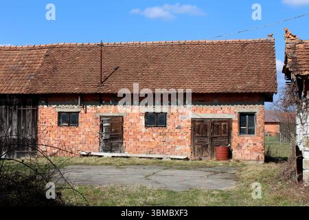 Rectangular outbuilding made of exposed red bricks features a steep tiled roof, two small windows, and weathered wooden doors, set in a grassy yard Stock Photo
