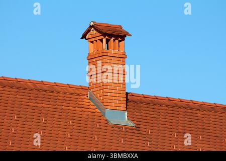 Tall red brick chimney with decorative cap rises from a red clay tile roof, with crisp flashing and regular tile pattern, set against a cloudless blue Stock Photo