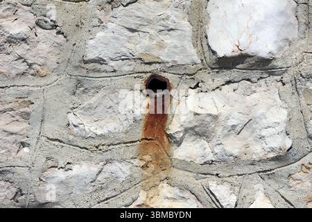 Weathered stone wall with a small rusty metal pipe protruding, leaving an orange-brown rust stain below, surrounded by rough light-colored stones Stock Photo