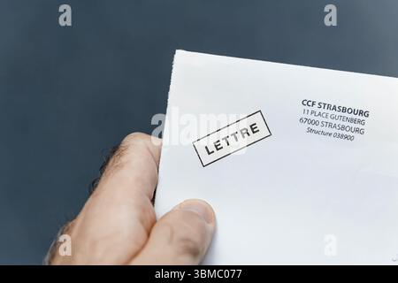 Paris, France - Oct 28, 2024: A hand holds a white envelope with the word LETTRE stamped on it and an address for CCF Strasbourg at 11 Place Gutenberg Stock Photo