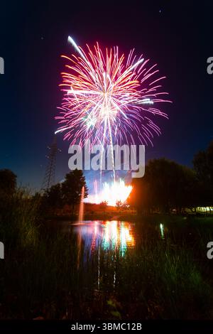 Bright fireworks explode overhead, reflecting on the calm water of a lake during a summer evening celebration. Stock Photo