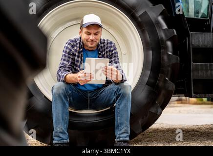 Modern agricultural worker sitting on tractor wheel and using digital tablet Stock Photo