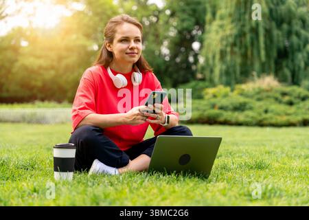 Young woman wearing red t-shirt using smart phone and laptop while sitting on grass in city park Stock Photo