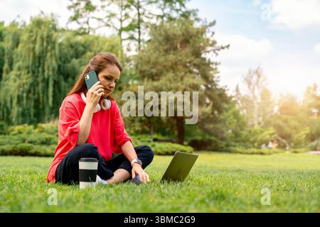 Young female freelancer using laptop and talking on phone outdoors Stock Photo