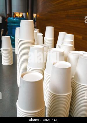 Stacks of white disposable paper cups arranged on a counter in an indoor space with people in the background and a wooden wall on the right side of th Stock Photo