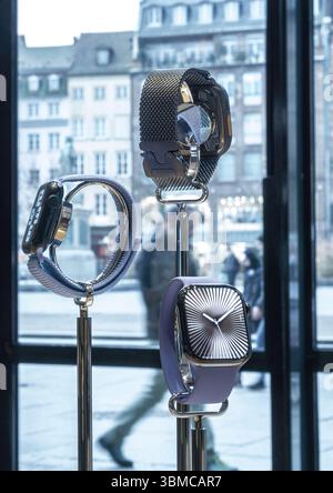 Paris, France - Mar 13, 2025: Stylish Apple Watches in diverse band styles are showcased on metal stands against a window revealing blurred pedestrians and traditional urban architecture outside Stock Photo