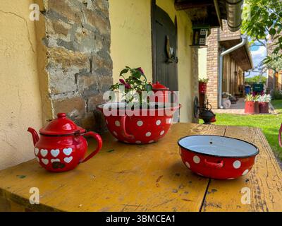 Old-fashioned red enamel teapot, bowl, and pot with white dots displayed on a wooden table in a charming rural courtyard, evoking nostalgia. Stock Photo