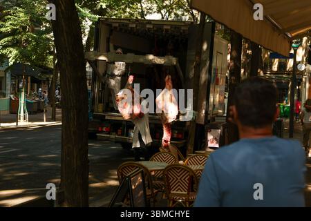 Two butchers in white coats unload large sides of beef from a refrigerated truck parked on a sunny Parisian street. Stock Photo