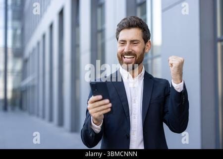 A happy businessman looks at his phone with a fist raised in the air, celebrating a success outdoors near a modern building. Stock Photo
