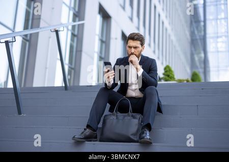 A concerned businessman in a suit is looking at his phone while sitting on steps in front of an office building. Stock Photo