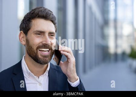 A smiling businessman in a suit, talking on a smartphone outdoors near a modern building. Stock Photo
