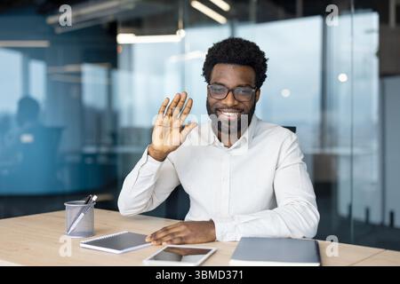 An African-American man in glasses waves and smiles while sitting at a desk in an office. He appears happy and approachable. Stock Photo