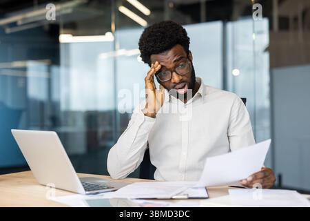 An African-American businessman is shocked while reviewing paperwork at his desk in the office. Stock Photo