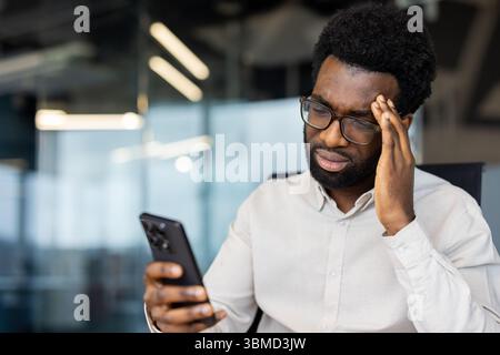 An African-American man with glasses looks at his phone, touching his forehead with a look of stress or concern in an office. Stock Photo