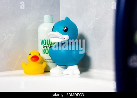 Two vibrant rubber ducks, one large blue and one small yellow, stand beside a light green shampoo bottle on the clean white edge of a bathtub. Stock Photo