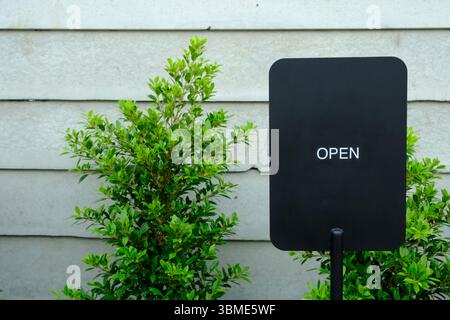 An outdoor OPEN sign is mounted on a textured wall next to lush green plants. This exterior view captures the business's readiness and accessibility f Stock Photo