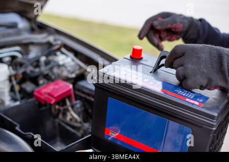 A man wearing gloves holds a battery pack over an engine bay, preparing to install it. Replacing a battery in a car. Stock Photo