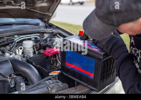 A man wearing gloves holds a battery pack over an engine bay, preparing to install it. Replacing a battery in a car. Stock Photo