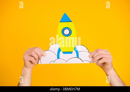 Bright yellow background showcasing hands holding a craft paper rocket with clouds representing creativity and imagination Stock Photo
