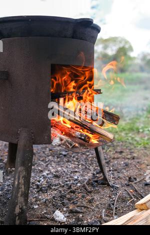 Fire Burns Brightly in a Rustic Metal Stove While Wood is Added for Cooking Outdoors in a Natural Setting During Daytime Stock Photo