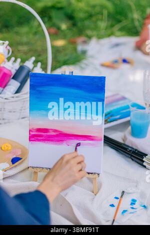 Outdoor art session showing woman's hand painting soft blue and magenta sky on canvas with mini easel under natural light. Stock Photo