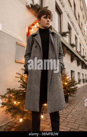 Handsome Pretty Young Man In A Elegant Gray Checkered Coat Is Standing Near The Building Between Two Christmas Beautiful Trees. European Guy Model Enj Stock Photo