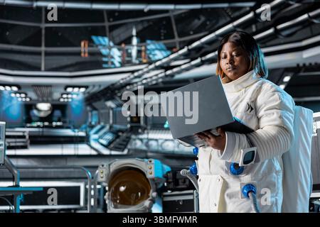 Astronaut using laptop to confirm suit integrity, monitoring life support systems. Orbital station crew member testing gear during routine equipment check using notebook, conducting safety checks Stock Photo