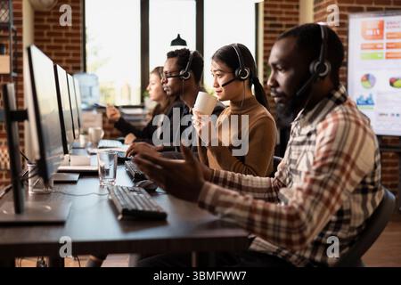 Asian female call center agent drinking cup of coffee and reviewing client information on her computer. Modern workspace with multiethnic telemarketers at work, speaking with online customers. Stock Photo