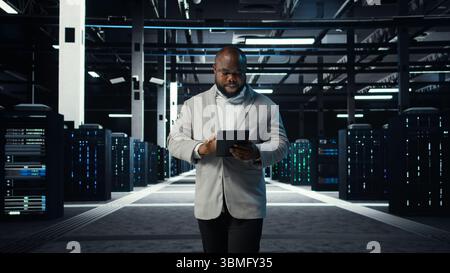 IT professional looking over data center checkup checklist paperwork on tablet. African american man walking in server farm, reading documentation files on device, camera A Stock Photo