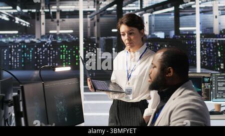 Happy data center manager providing positive feedback to engineer developing neural networks software on computer. Cheerful supervisor pleased by employee work implementing AI in server room, camera A Stock Photo
