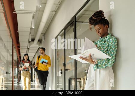 Young adult Black woman standing in hallway reading documents while colleagues walking together in background holding notebooks and folders Stock Photo