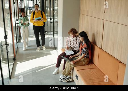 Young Caucasian man and Asian woman sitting on bench collaborating with laptop while Black colleagues walking in modern coworking hallway Stock Photo