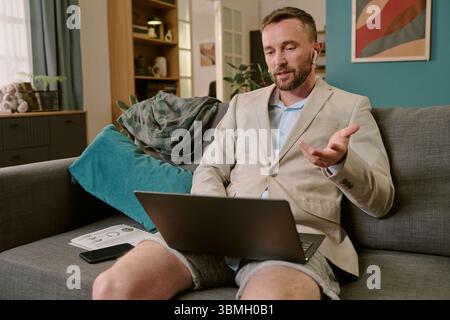 Young Adult Man Working Remotely Using Laptop Sitting on Sofa Stock Photo
