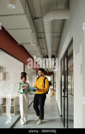 Black woman holding notebook talking with man wearing glasses and yellow sweatshirt carrying laptop and backpack in modern coworking hallway Stock Photo