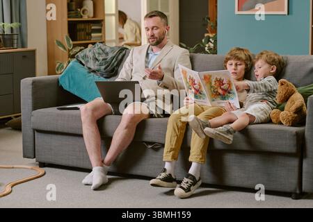 Middle Aged Man Working Remotely While Sons Reading Storybook Stock Photo
