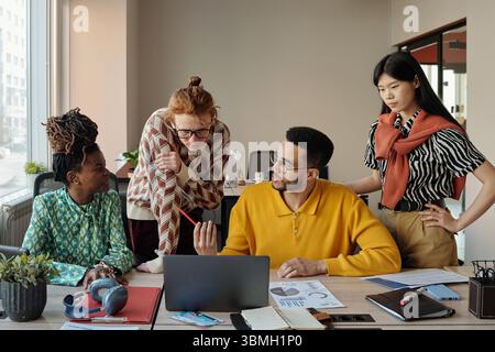 Young adult diverse men and women collaborating around laptop in modern coworking space, discussing project documents Stock Photo
