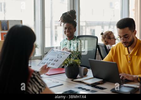 Young adult Black woman smiling while discussing business charts with colleague across desk, young adult man working on laptop Stock Photo