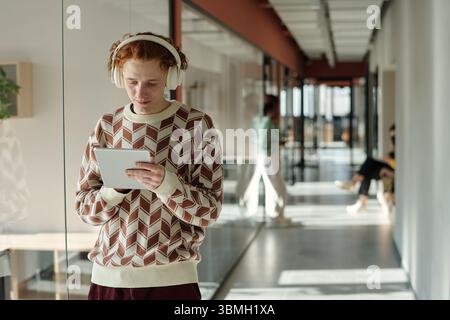 Young Caucasian man with red hair wearing patterned sweater and headphones standing in modern coworking space using digital tablet Stock Photo