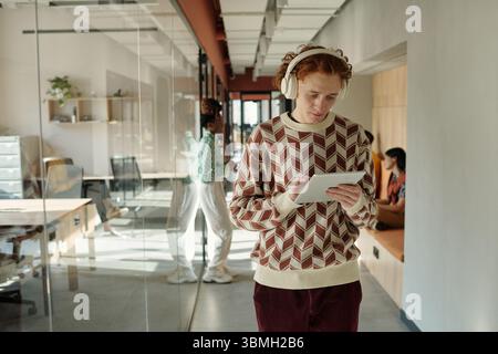 Caucasian man with red hair wearing headphones using digital tablet while walking in modern coworking space, two women collaborating in background Stock Photo
