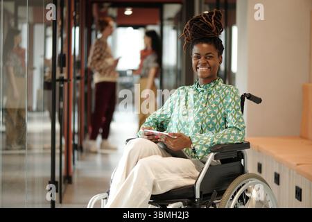 Black woman sitting in wheelchair holding digital tablet smiling at camera in coworking space with two young adult colleagues talking in background Stock Photo