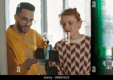 Young Caucasian man with red hair and Black man with glasses collaborating on creative coworking project, arranging sticky notes on glass wall Stock Photo