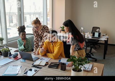 Gen Z diverse people collaborating around desk with laptop and documents in modern coworking office, discussing project Stock Photo