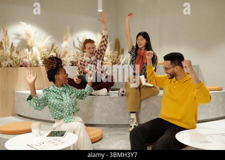 Group of young adult coworkers sitting on steps in creative coworking space raising hands and smiling, holding digital tablets and papers Stock Photo