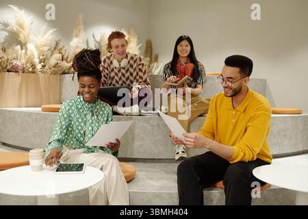 Group of young adult coworkers sitting on steps in creative coworking space holding papers and digital tablet, smiling Stock Photo